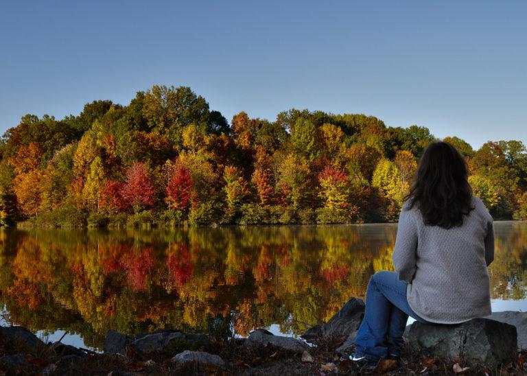 woman looking across lake at autum trees which are reflecting in the water